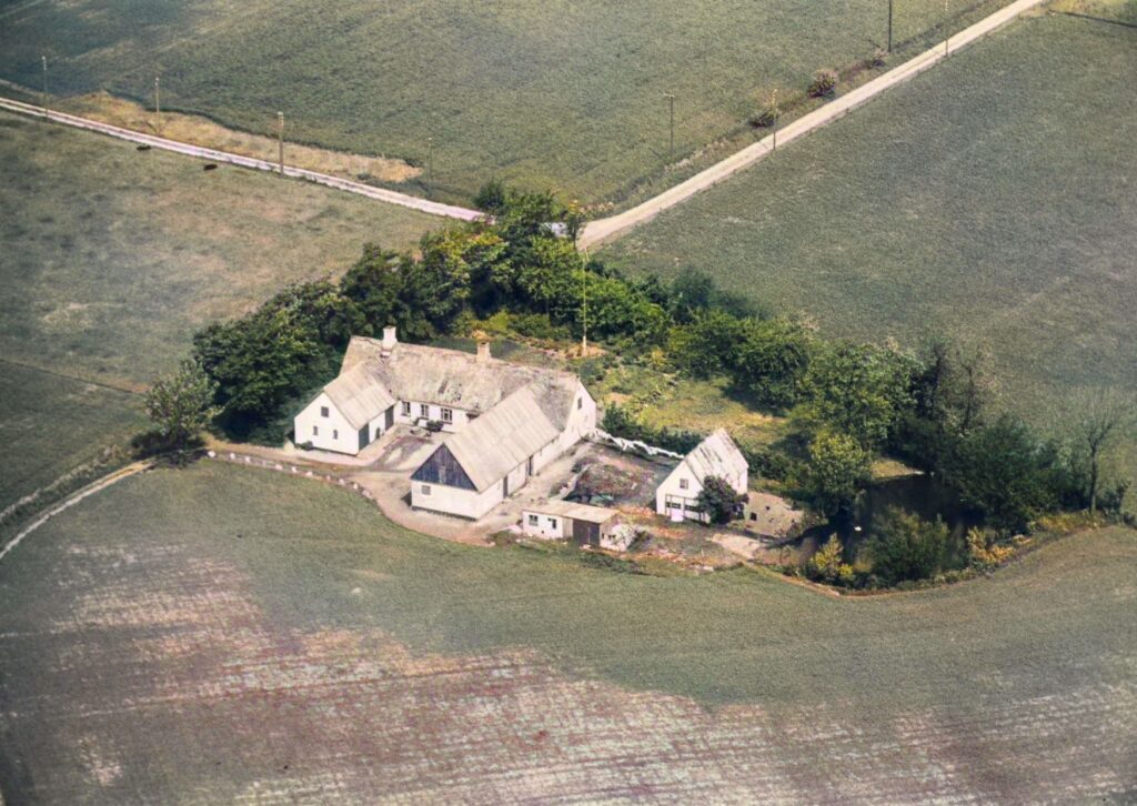 En 3-længet bondegård med nedslidt stråtag. Bag gården et udhus og et mindre hus ved siden af en dam.